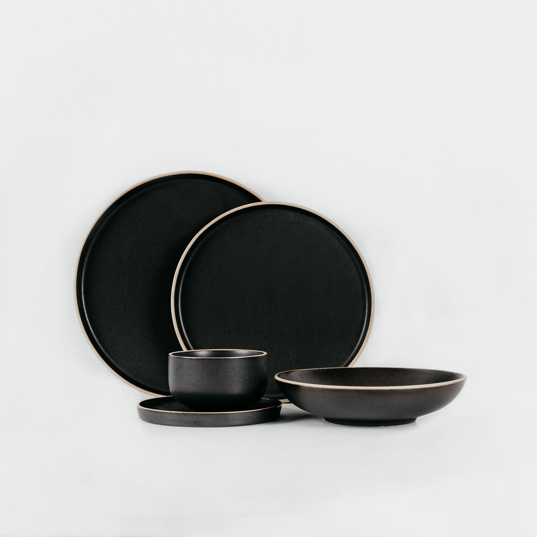 A set of matte black ceramic tableware, including two large plates, a small bowl with its saucer, and a shallow bowl, arranged on a clean white background.