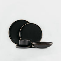 A set of matte black ceramic tableware, including two large plates, a small bowl with its saucer, and a shallow bowl, arranged on a clean white background.